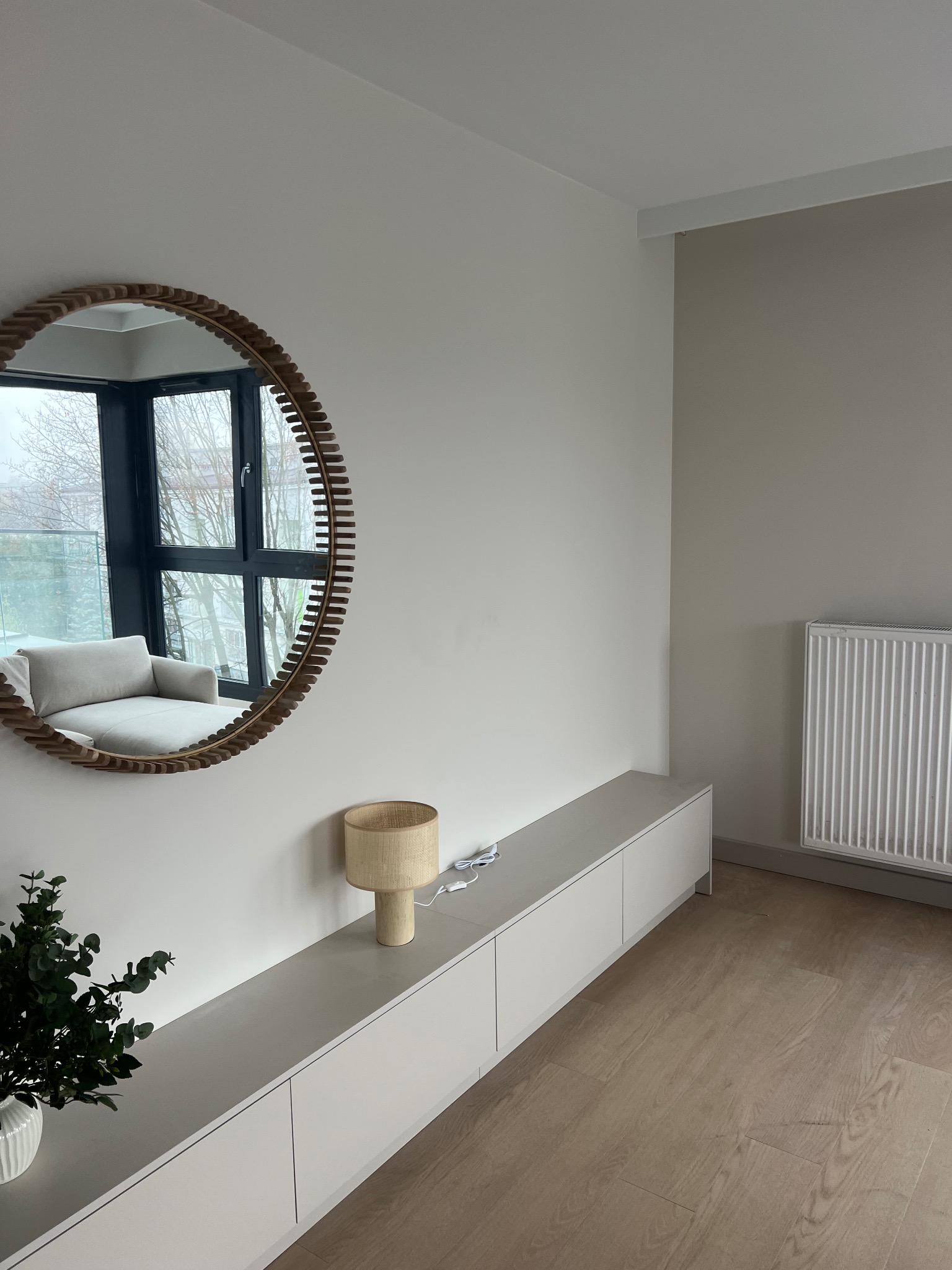Jasne wnętrze z okrągłym lustrem z drewnianą ramą, podłużną szafką RTV, lampką i grzejnikiem. Minimalistyczny design, parkiet na podłodze.