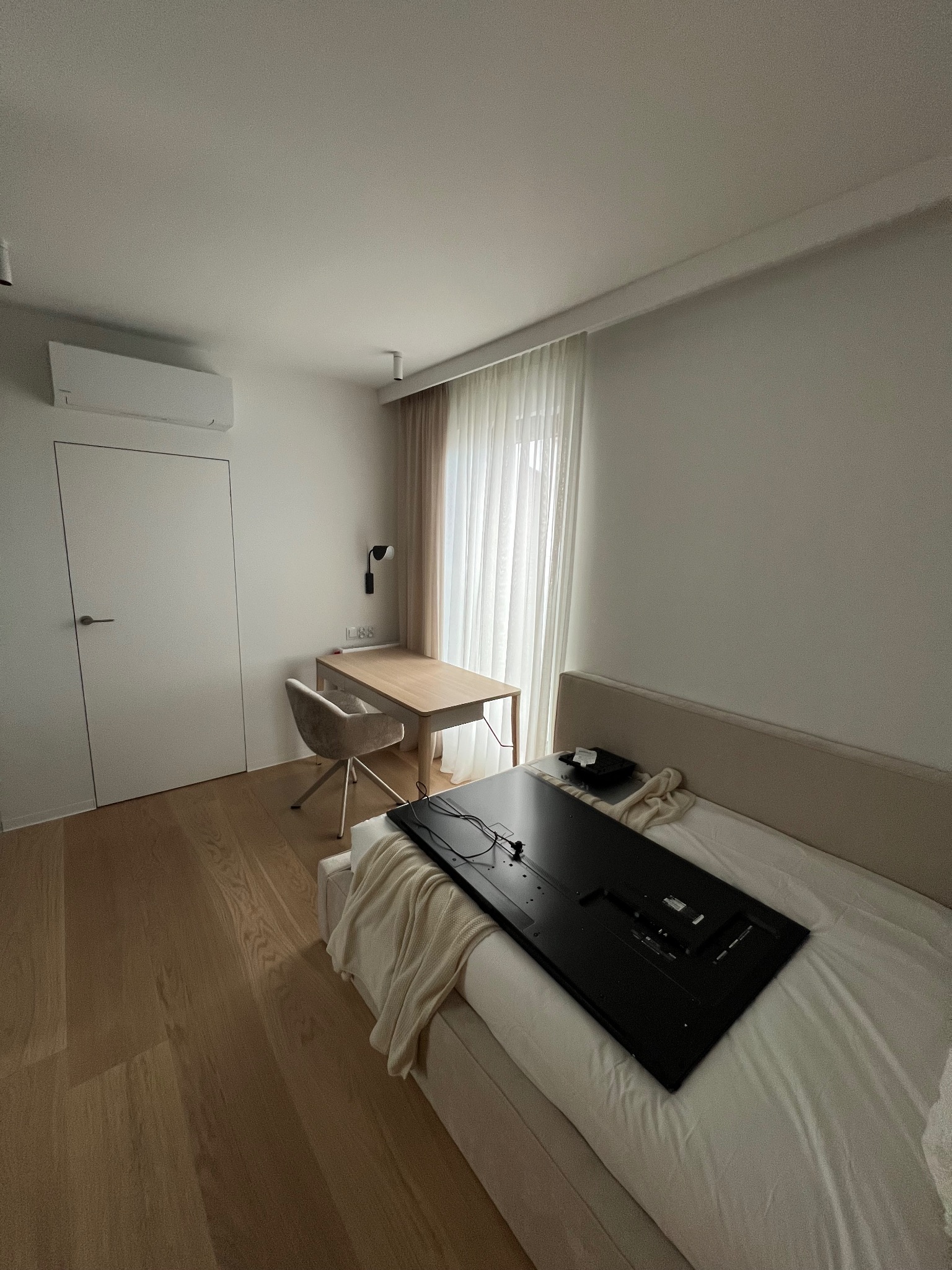 Minimalistyczne wnętrze pokoju z drewnianą podłogą, białymi ścianami i meblami. Na łóżku leży duży telewizor, obok biurko z krzesłem i zasłonięte okno.