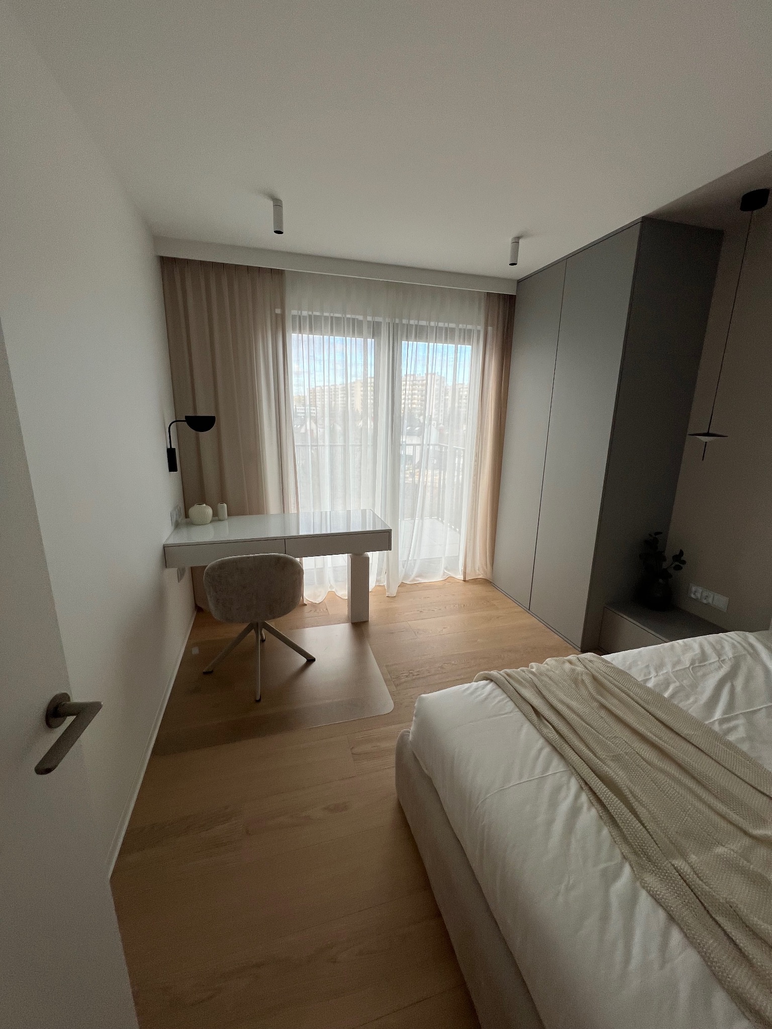 Jasne wnętrze sypialni z biurkiem, krzesłem i łóżkiem. Parkiet na podłodze, okno z firankami w tle. Minimalistyczny design, stonowana kolorystyka.