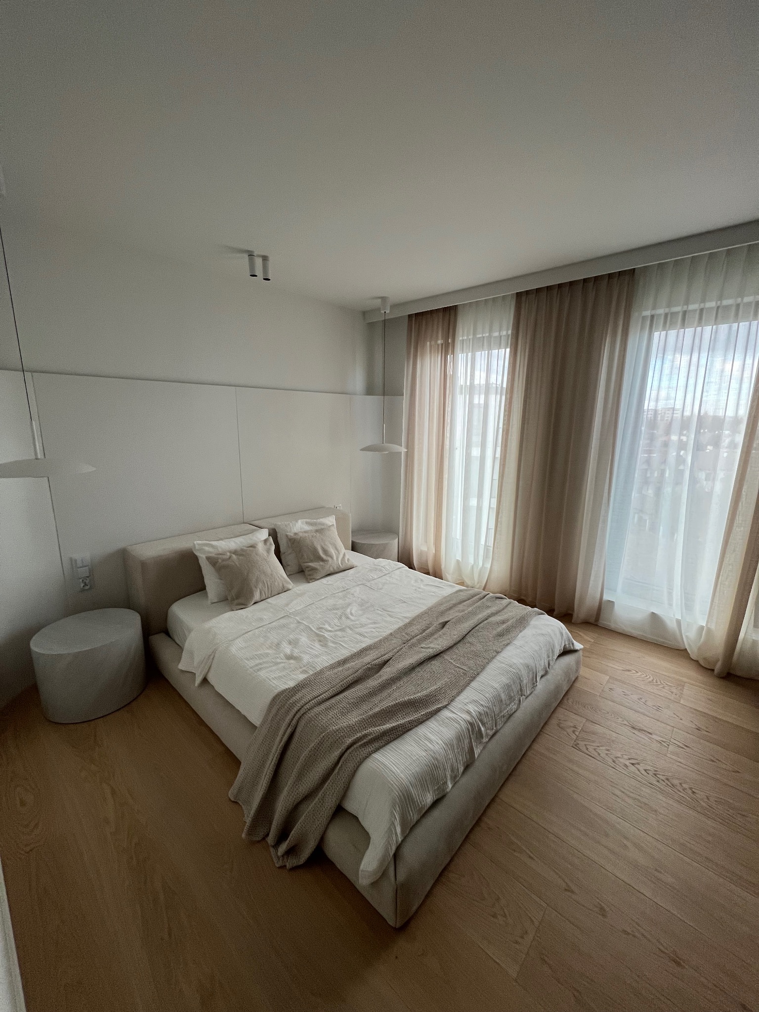 Jasna sypialnia z drewnianą podłogą, beżowym łóżkiem i zasłonami. Minimalistyczny wystrój, stonowana kolorystyka, widok na miasto za oknem. Nowoczesne lampy wiszące.