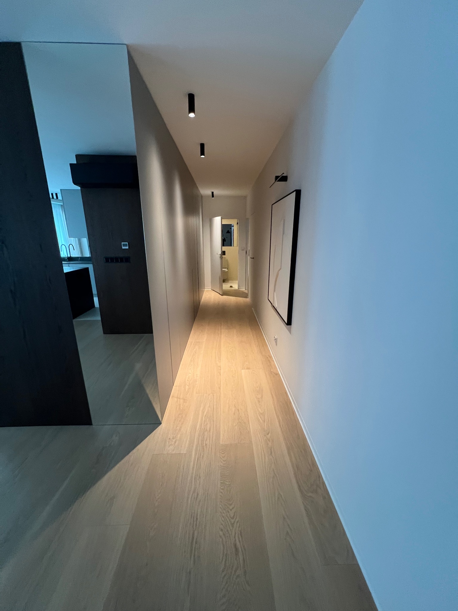 Długi korytarz z drewnianą podłogą i minimalistycznym oświetleniem punktowym. Po lewej stronie lustro i zabudowa, po prawej biała ściana z obrazem w czarnej ramie.