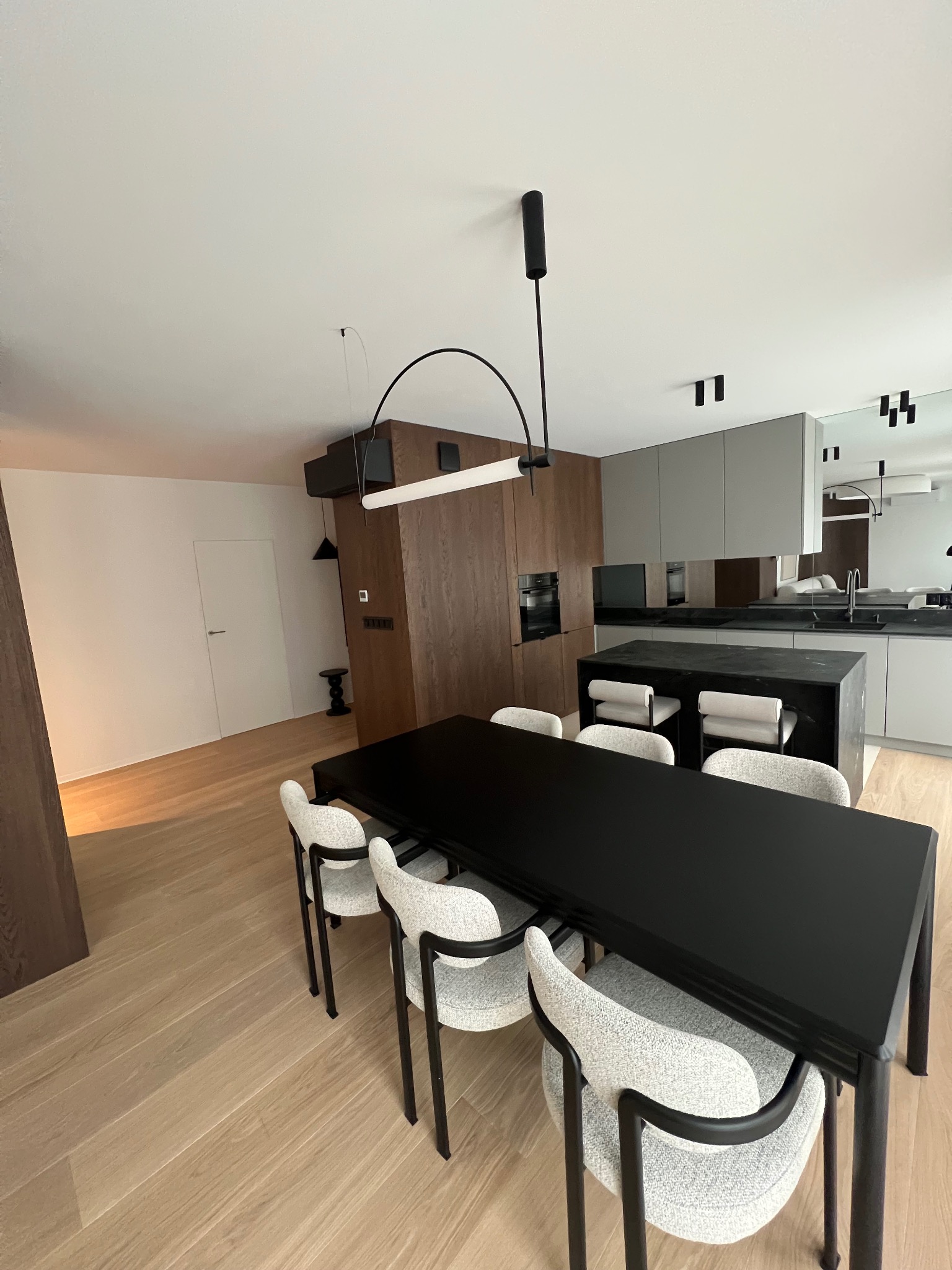 Nowoczesne wnętrze: drewniana zabudowa, szara kuchnia z wyspą i czarny stół z krzesłami na jasnej podłodze. Minimalistyczne oświetlenie.