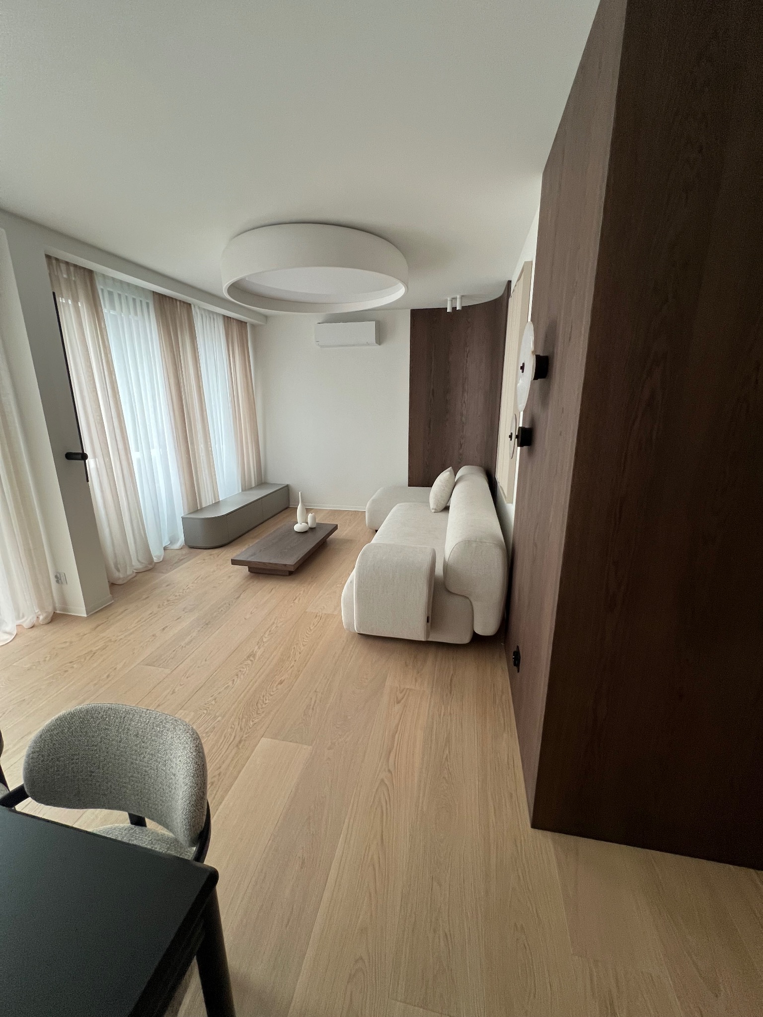 Jasne, przestronne wnętrze salonu z drewnianą podłogą, beżową sofą i minimalistycznym oświetleniem. Ciemna, drewniana ściana dodaje kontrastu. Widok na okno z delikatnymi zasłonami.