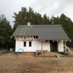 Budowa domu murowanego: widok na bryłę budynku z białych bloczków, z częściowo ukończonym dachem pokrytym papą i zielonymi łatami, na tle lasu.