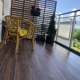 Balkon z brązowymi płytkami imitującymi drewno, rattanowym fotelem z żółtą poduszką, drewnianą osłoną, kwiatami w doniczkach i szklaną balustradą z widokiem na zieleń.