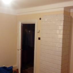 KSAPAT - Świeżo pomalowane ściany w pokoju z białym, kaflowym piecem narożnym, widoczna drabina malarska i zabezpieczona folią podłoga przy ścianie.