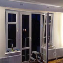 KSAPAT - Białe, przeszklone drzwi balkonowe w trakcie montażu, częściowo otwarte, widoczna puszka silikonu i drabina, ślady prac remontowych.