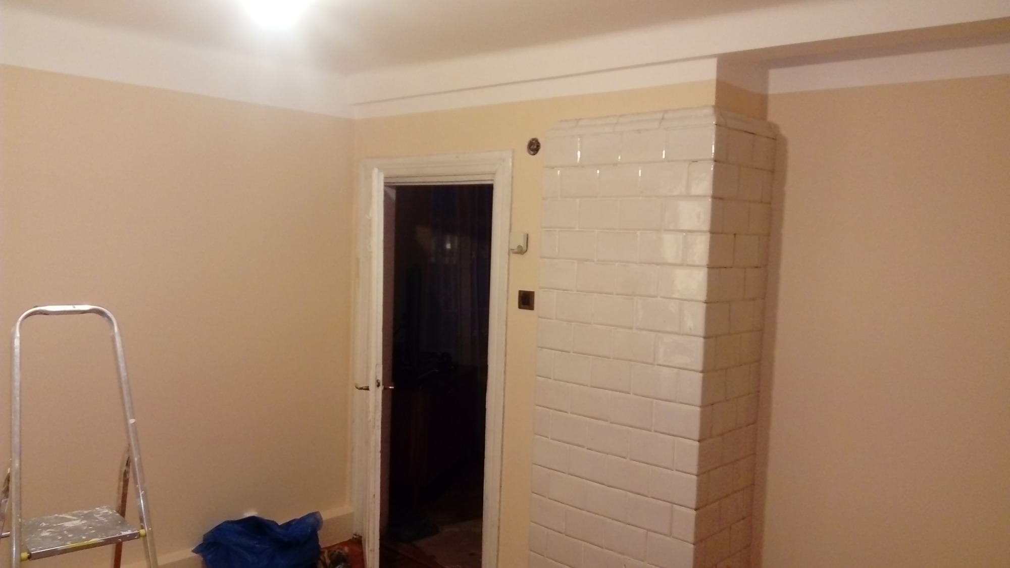 Świeżo pomalowane ściany w pokoju z białym, kaflowym piecem narożnym, widoczna drabina malarska i zabezpieczona folią podłoga przy ścianie.