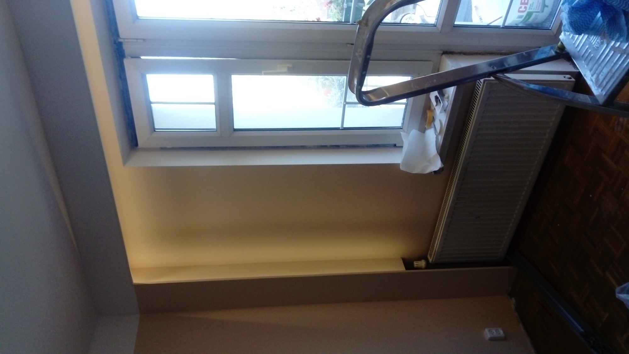 Świeżo pomalowana ściana przy oknie z grzejnikiem, widoczna drabina malarska i zabezpieczenia.