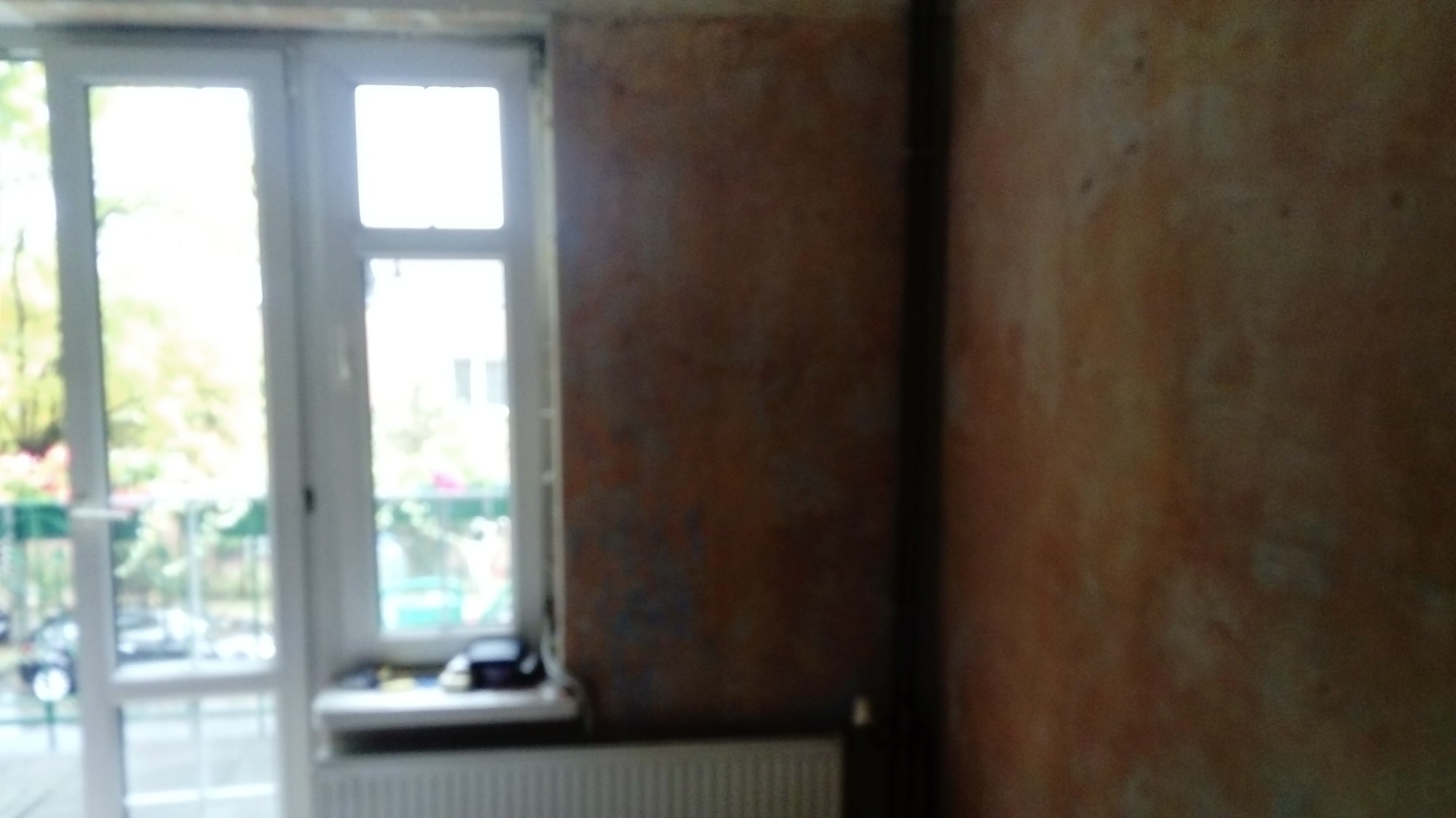 Ściana z nierówną, pomarańczowo-brązową fakturą przed malowaniem, widoczne fragmenty starej farby, okno balkonowe z widokiem na zieleń w tle.