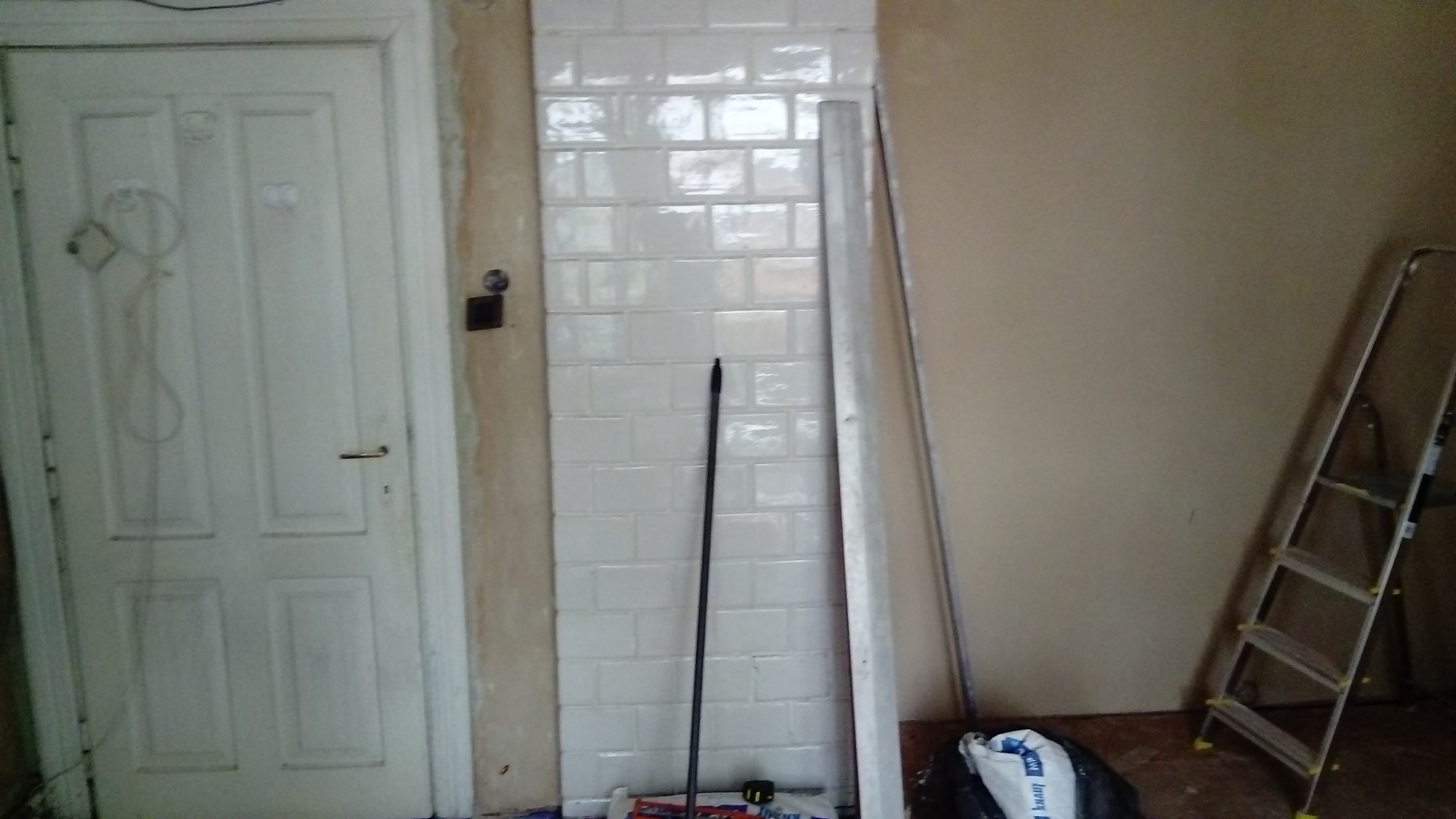 Fragment ściany wyłożony białymi, błyszczącymi kaflami typu metro, widoczna drabina, białe drzwi z ościeżnicą w trakcie prac remontowych, oraz worek z klejem.