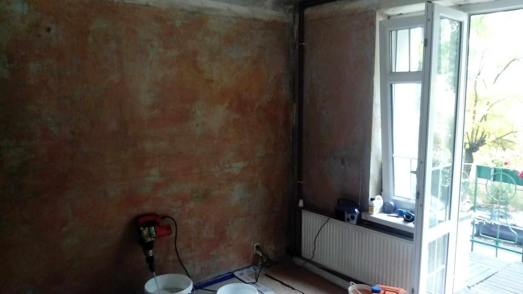 Ściana w trakcie malowania dekoracyjnego z widocznymi smugami farby w odcieniach brązu i beżu, mieszadło do farb w białym wiadrze, otwarte okno balkonowe z widokiem na zieleń.