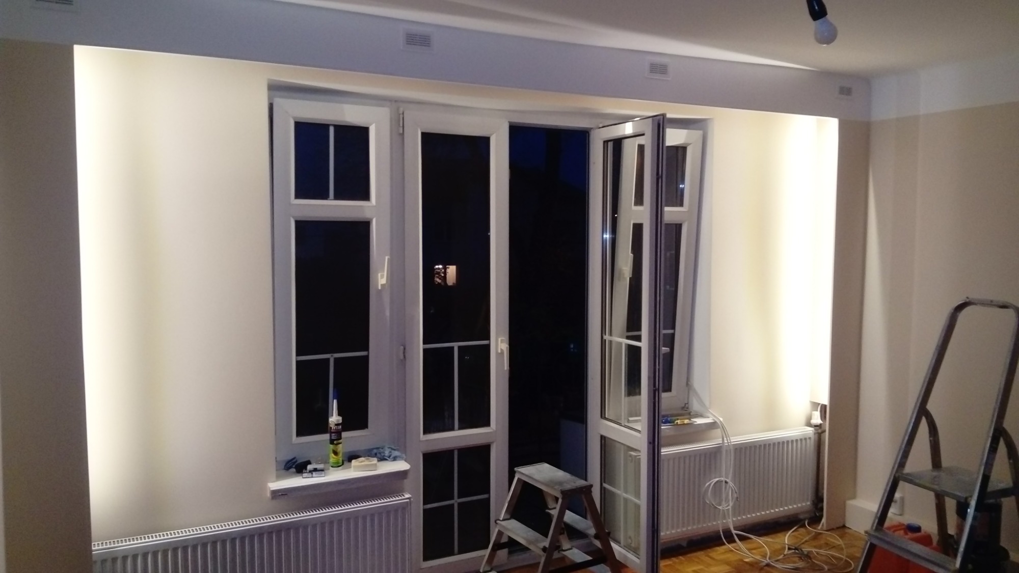 Białe, przeszklone drzwi balkonowe w trakcie montażu, częściowo otwarte, widoczna puszka silikonu i drabina, ślady prac remontowych.