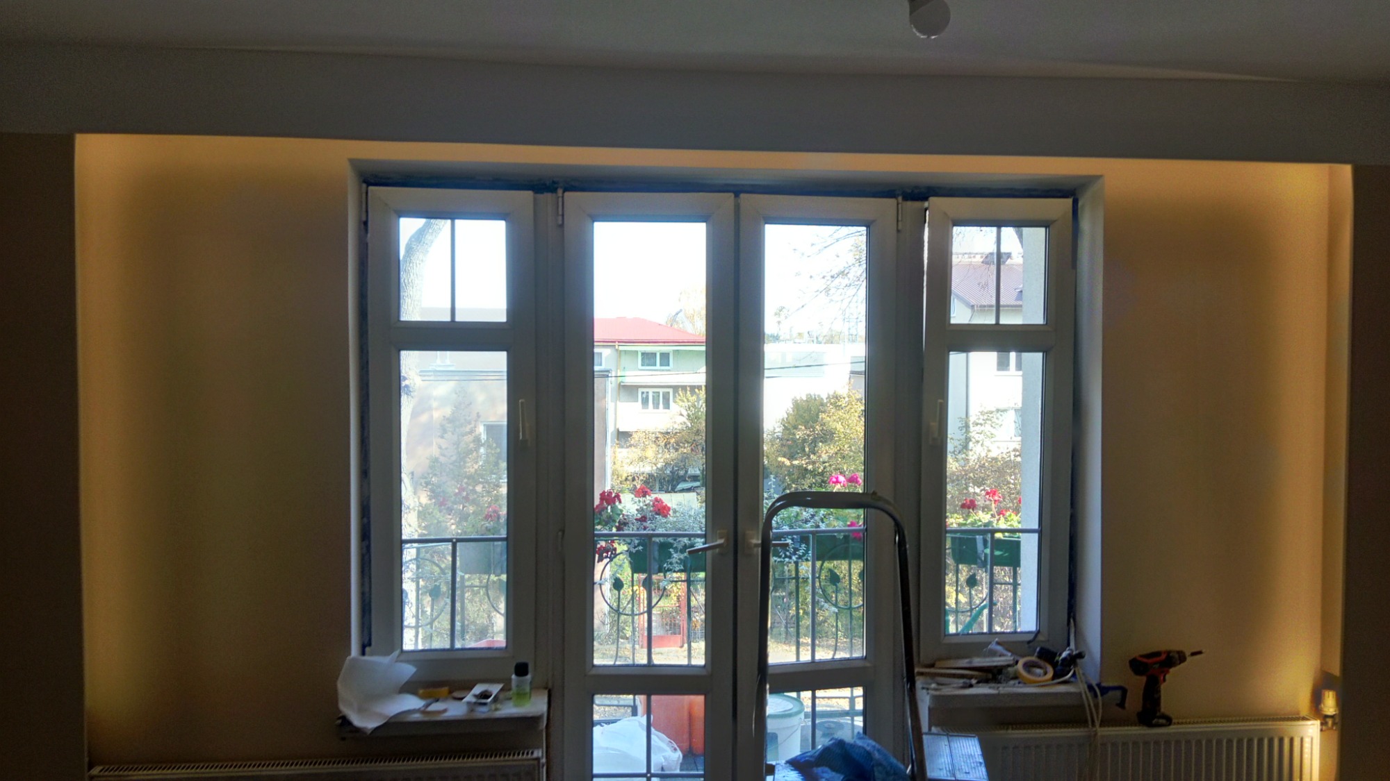 Wnętrze pokoju z dużym oknem balkonowym, drabina oparta o okno, widok na balkon z kwiatami, ślady po taśmie malarskiej na ramie okna, narzędzia na parapecie, przygotowania do malowania ścian.