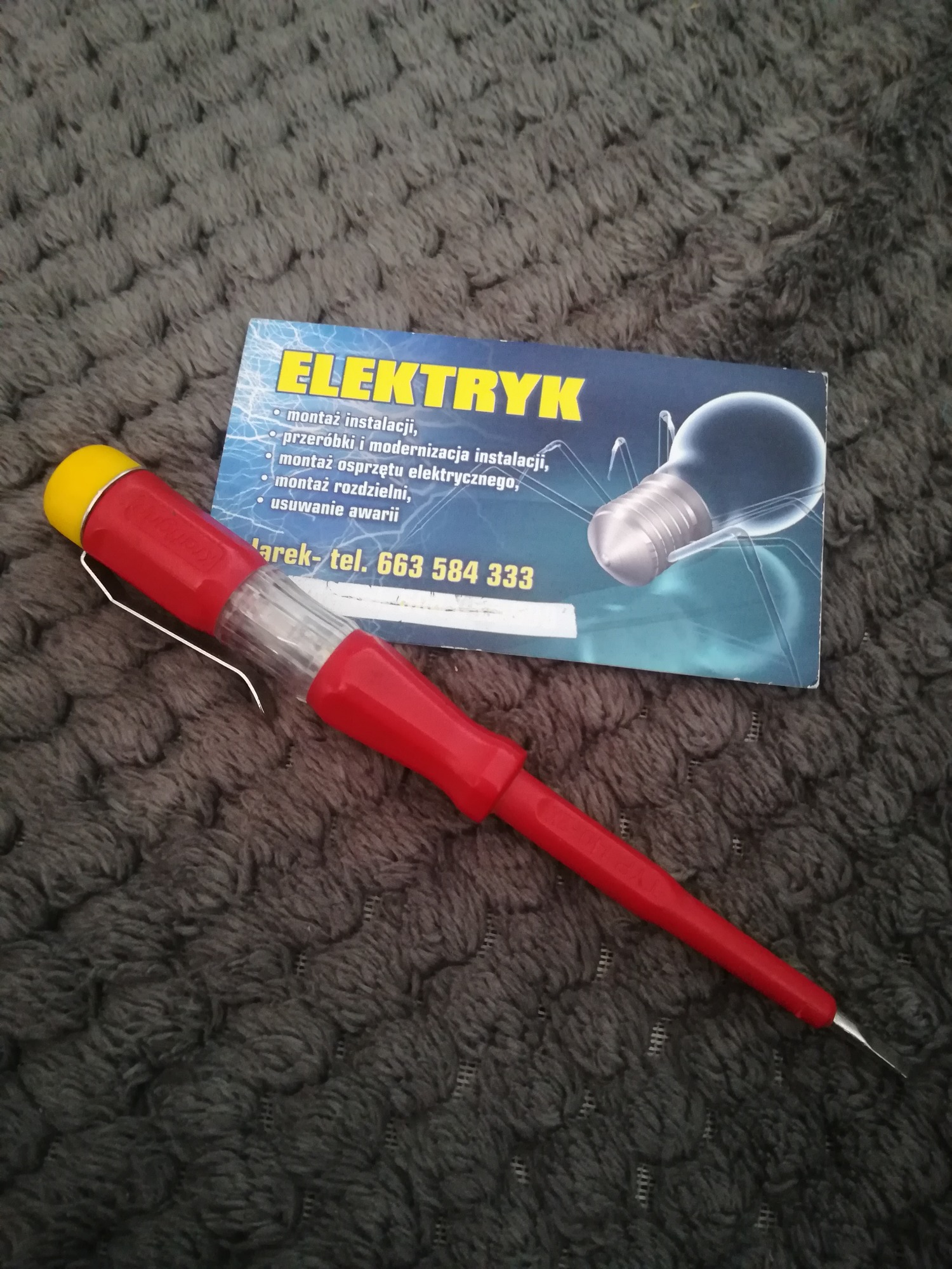 Wskaźnik napięcia z czerwoną rączką i żółtą końcówką leżący obok wizytówki z napisem ELEKTRYK na tle szarej, puszystej tkaniny.