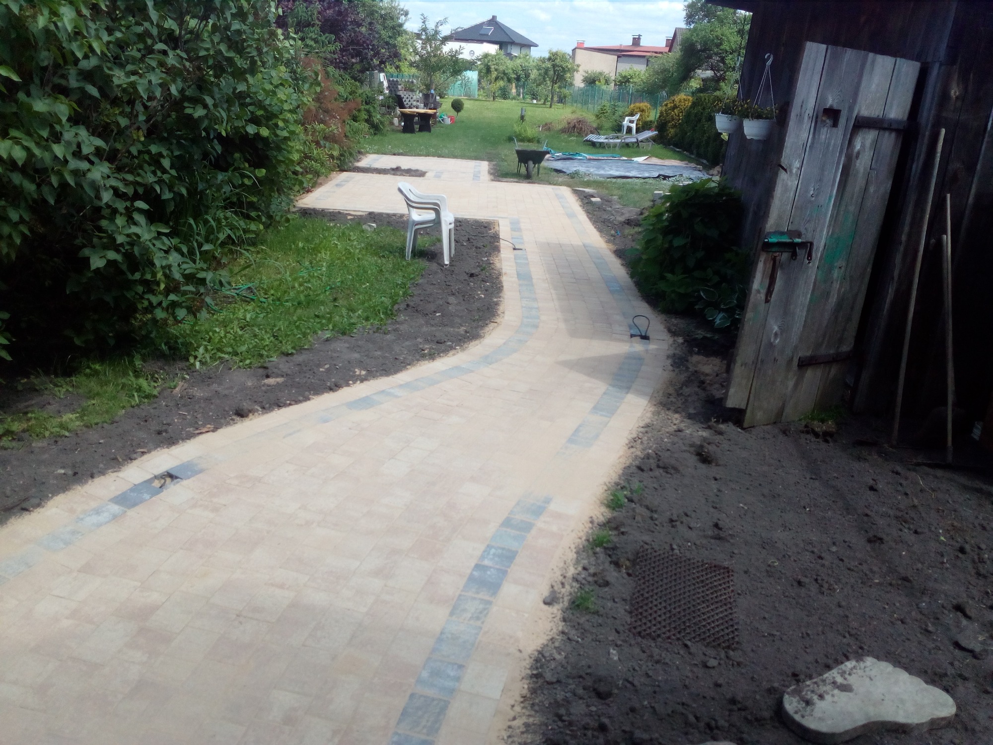 Kremowa ścieżka z kostki brukowej z szarymi wstawkami wijąca się przez ogród, widok z poziomu gruntu w kierunku domu, częściowo zarośnięte krawędzie, białe krzesło ogrodowe po lewej stronie.