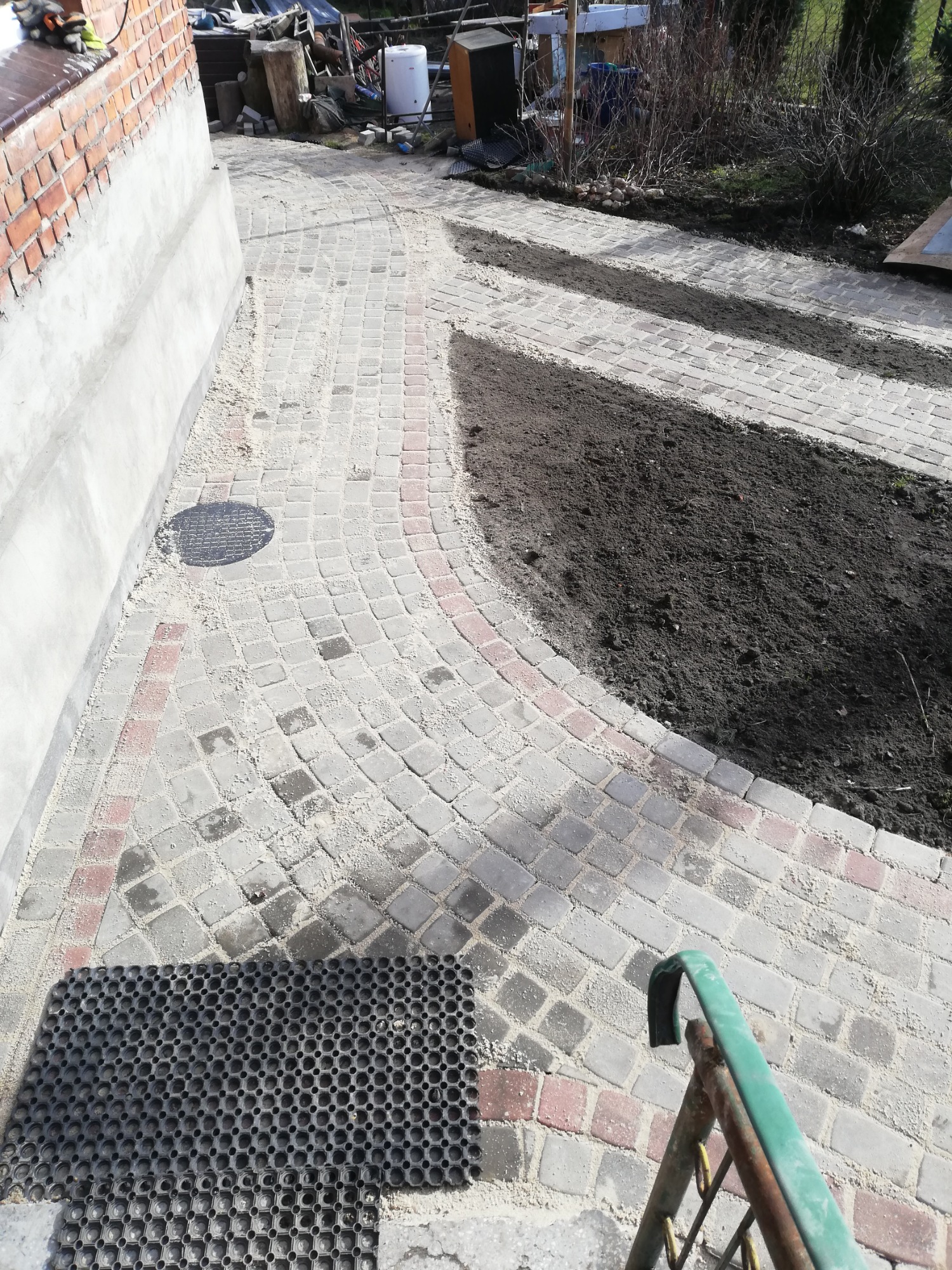 Zakrzywiony chodnik z kostki brukowej w odcieniach szarości i czerwieni, z czarną wycieraczką w rogu, otoczony przygotowanym gruntem pod nasadzenia, widok z góry.