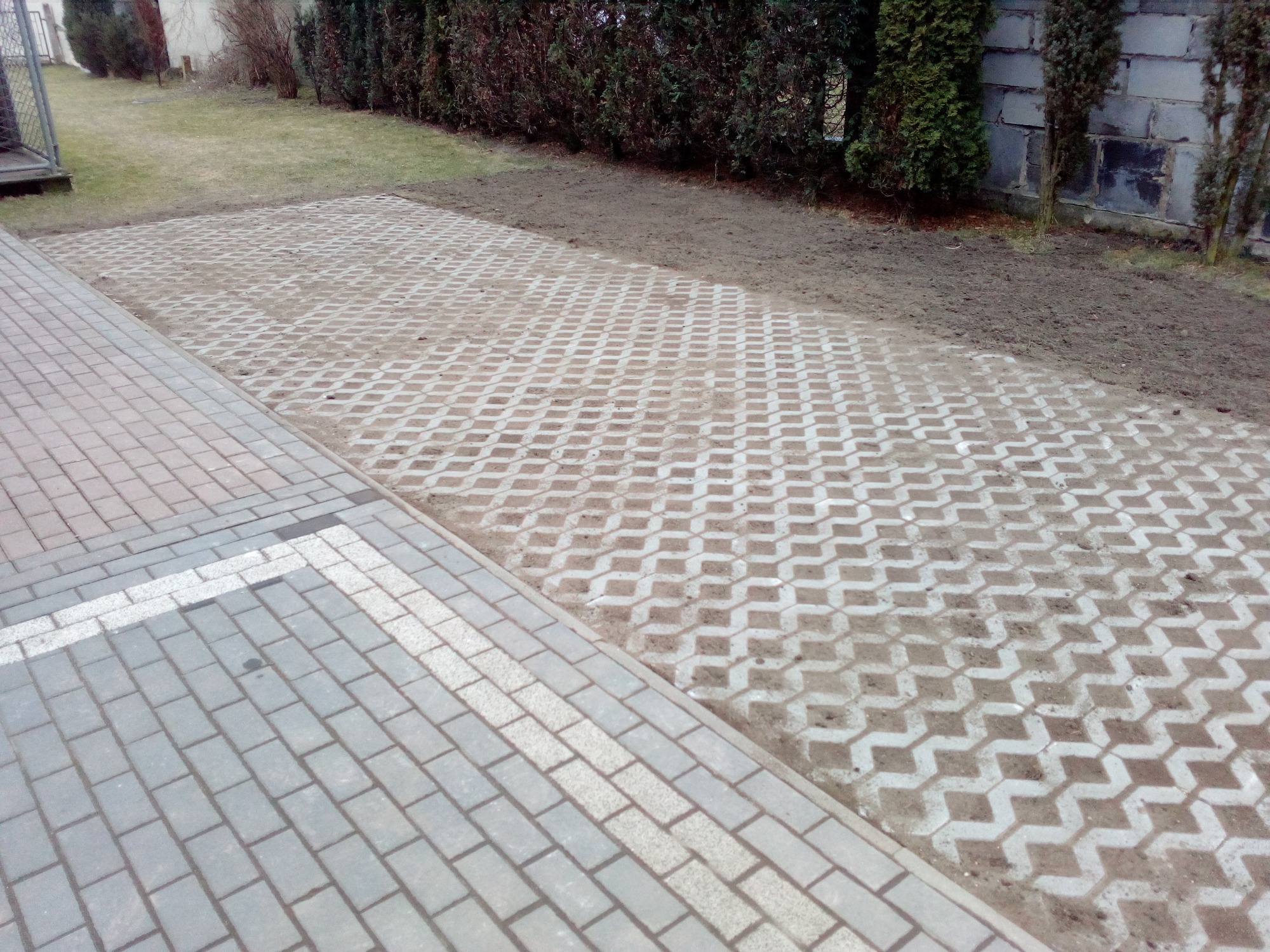 Ułożona nawierzchnia z ażurowych płyt betonowych wypełnionych ziemią, sąsiadująca z chodnikiem z szarej kostki brukowej z białym pasem, obrzeża z ziemi, w tle żywopłot i budynek.