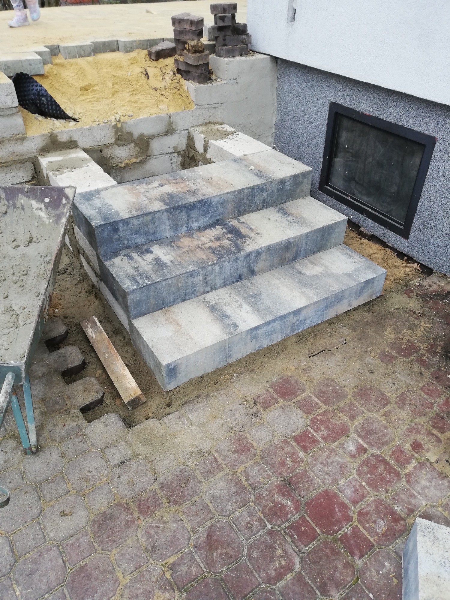 Budowa schodów zewnętrznych z kostki brukowej o szarym, cieniowanym wzorze, widoczna taczka z zaprawą i przygotowane podłoże z piasku.