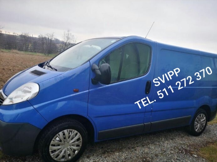 Niebieski bus dostawczy Opel Vivaro z numerem telefonu SVIPP 511 272 370 na boku, zaparkowany na żwirowym podłożu, w tle zaorane pole i pochmurne niebo.