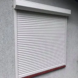 Zamknięta, biała roleta zewnętrzna na oknie budynku z szarą, chropowatą elewacją. Widoczny fragment brązowego parapetu i drewnianej podłogi przy ścianie.