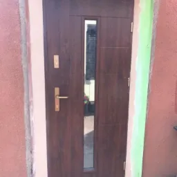 Brązowe drzwi zewnętrzne z długim, pionowym przeszkleniem, widoczne ślady po montażu na ościeżnicy i ścianie zewnętrznej budynku.