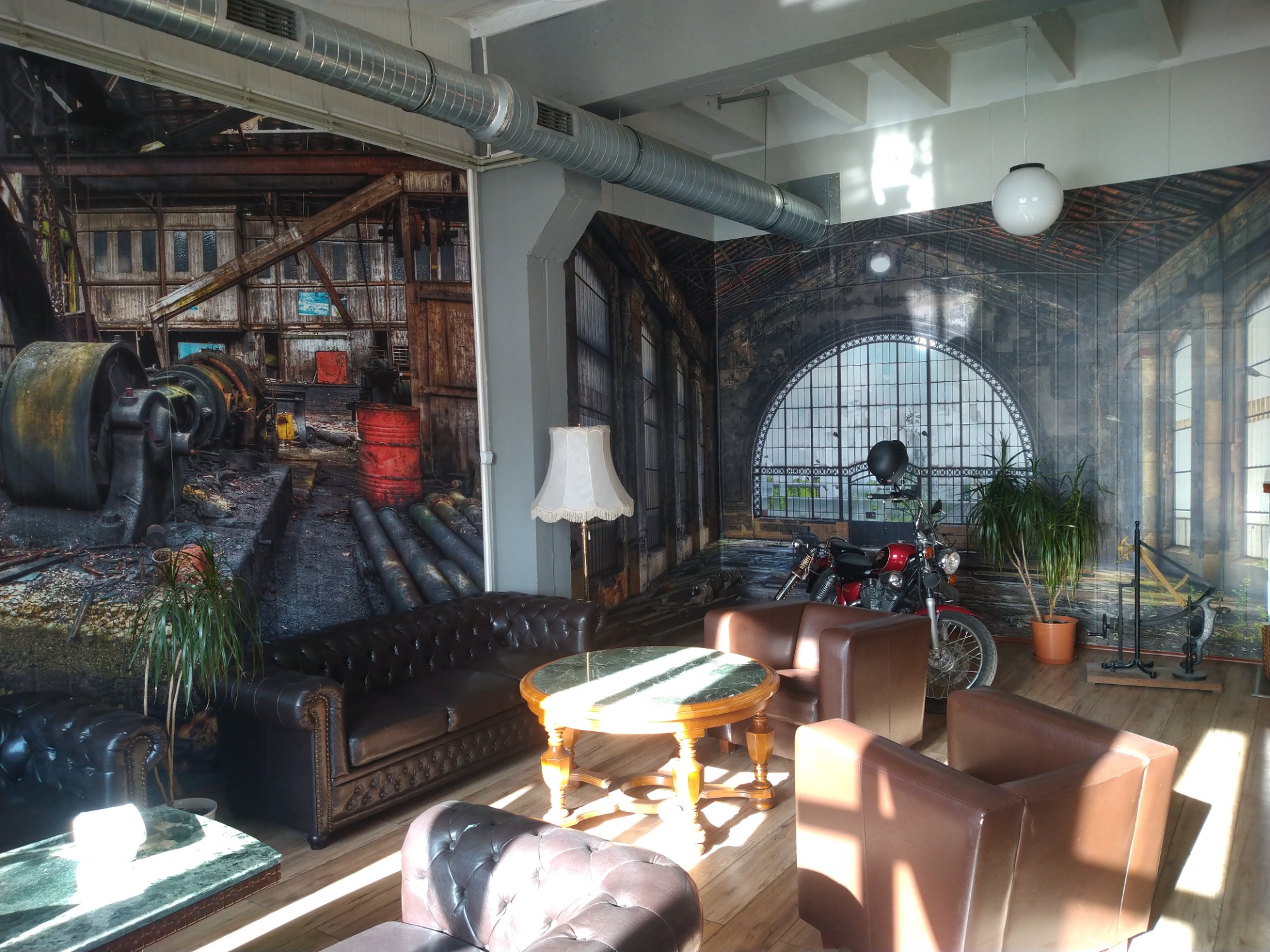 Wnętrze kawiarni lub restauracji z fototapetami przedstawiającymi industrialne sceny, skórzane sofy Chesterfield, okrągły stolik z zielonym blatem, czerwony motocykl i lampa podłogowa z abażurem.