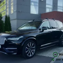Czarny SUV Volvo XC90 z relingami dachowymi zaparkowany przed nowoczesnym budynkiem biurowym z zielonymi tujami.