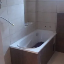 Wykończona łazienka z obudowaną wanną w brązowe panele, białe płytki na ścianach i wnęka w ścianie z półką.