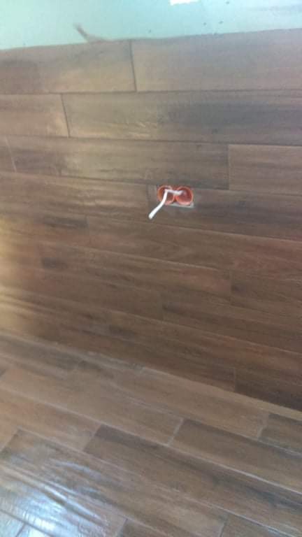 Ułożone poziomo brązowe płytki imitujące drewno na ścianie, z wystającym pomarańczowym gniazdkiem elektrycznym i białymi kablami.