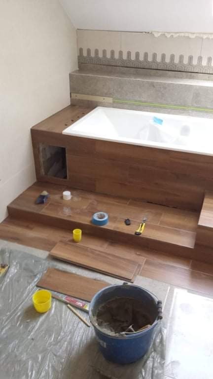 Montaż płytek imitujących drewno wokół wanny, widoczne narzędzia i zaprawa w niebieskim wiadrze, częściowo wykończona ściana z ozdobnym paskiem płytek.