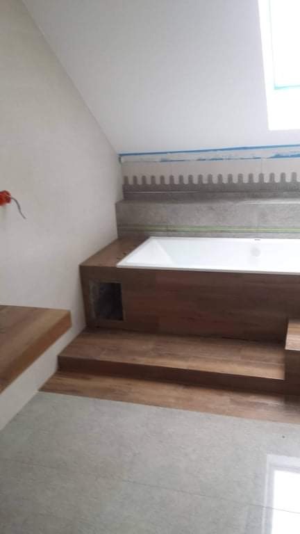 Wanna obudowana panelami imitującymi drewno, z widocznym otworem rewizyjnym, na tle ściany z szarą mozaiką pod sufitem w skosie.