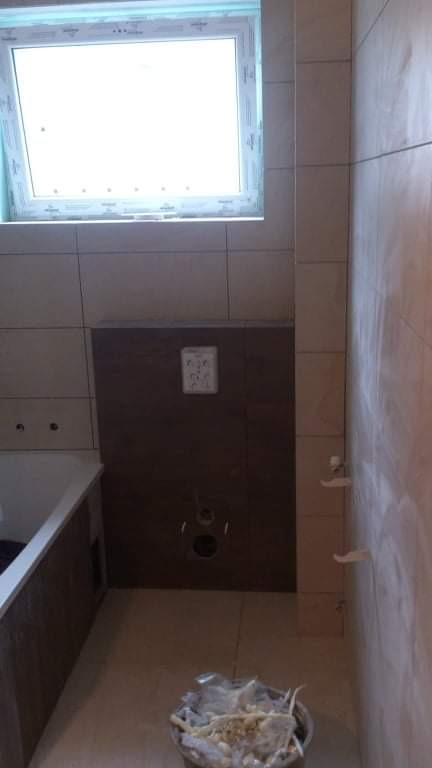 Wnętrze łazienki w trakcie remontu w Katowicach, z widoczną częściowo wykafelkowaną ścianą, wanną i oknem, oraz wiadrem z odpadami budowlanymi na podłodze.