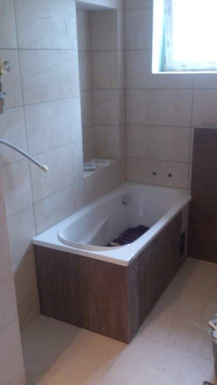 Wykończona łazienka z obudowaną wanną w brązowe panele, białe płytki na ścianach i wnęka w ścianie z półką.