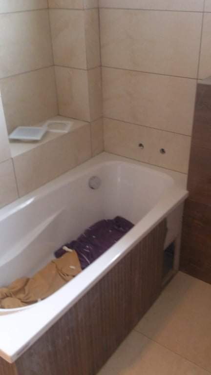 Wykończona łazienka z beżowymi płytkami i białą wanną z obudową imitującą drewno; widoczne otwory na baterię i akcesoria łazienkowe; w wannie leżą ubrania.