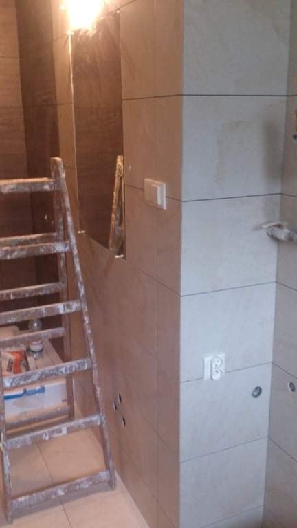 Ściana łazienki w trakcie wykańczania płytkami w odcieniach beżu i brązu, widoczna drabina malarska z resztkami zaprawy i otwory pod gniazdka elektryczne.