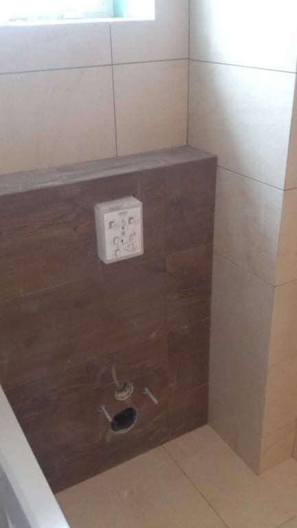 Fragment łazienki w trakcie remontu: brązowe płytki imitujące drewno na zabudowie podtynkowego stelaża WC, widoczne otwory na rury i przycisk spłuczki, jasne płytki na ścianach i podłodze.