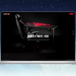Laptop wyświetlający reklamę biurka i krzesła gamingowego Diablo X-Mate 1400 na czarnym tle, z czerwonym motywem graficznym na biurku i krześle. Widoczny fragment tekstu reklamowego o zapewnieniu...