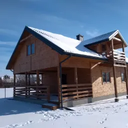 Zimowy krajobraz z nowo wybudowanym, drewnianym domem szkieletowym z balkonem i śniegiem na dachu, widoczne ślady na śniegu i samochód w tle.