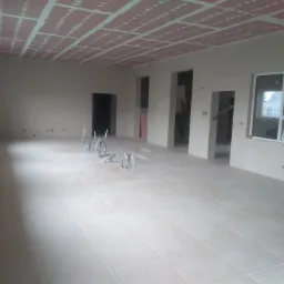 Wnętrze w trakcie remontu: ułożona jasna podłoga z płytek, sufit z płyt karton-gips z widocznymi łączeniami, surowe ściany z otworami na drzwi i okna, widoczne metalowe konstrukcje na podłodze.