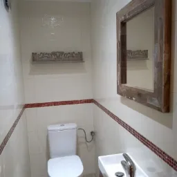 Niewielka łazienka po remoncie z toaletą, umywalką nablatową z chromowaną baterią, lustrem w drewnianej ramie i dekoracyjną półką na białych płytkach z bordiurą.