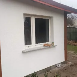 Biała elewacja domu z oknem i kamiennym parapetem, na którym leżą przekąski i słoik, obok drewniana ściana i stół w ogrodzie.