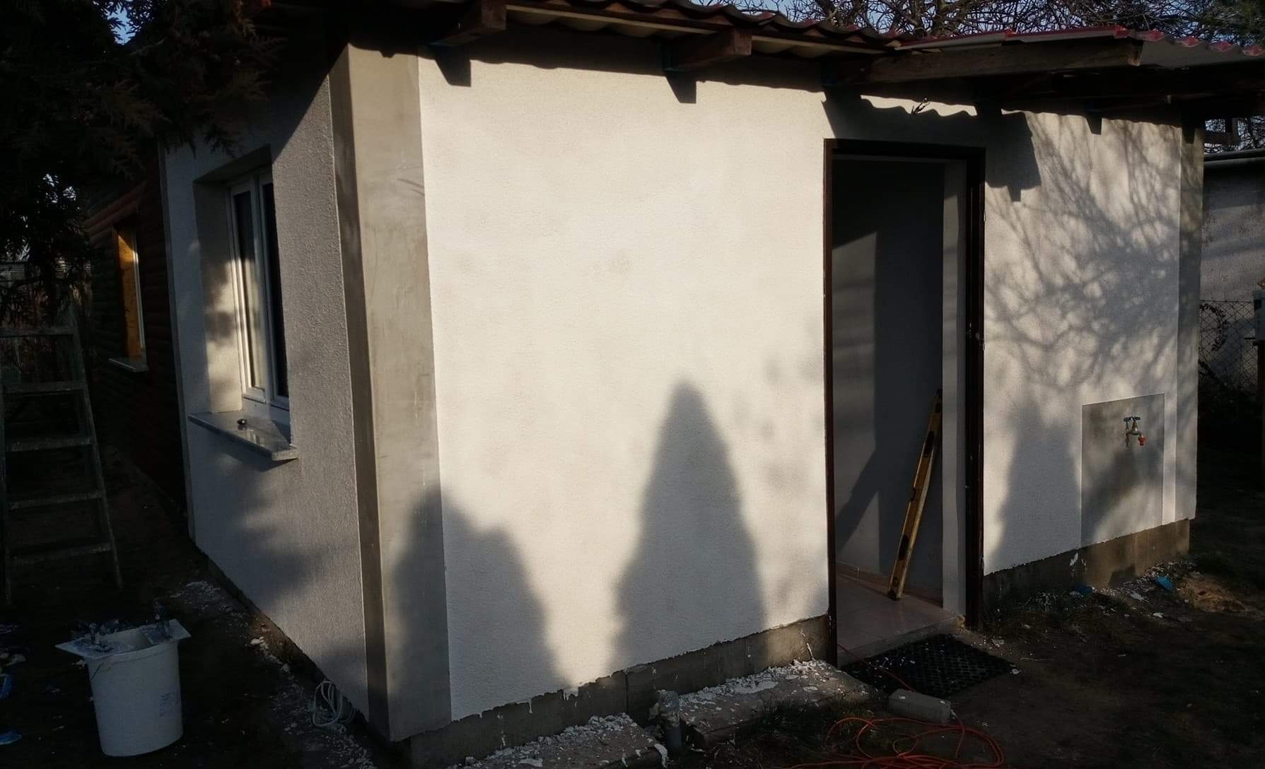 Otynkowana ściana budynku gospodarczego z widocznym wejściem, fragmentem dachu, oknem i drzewami rzucającymi cień na elewację