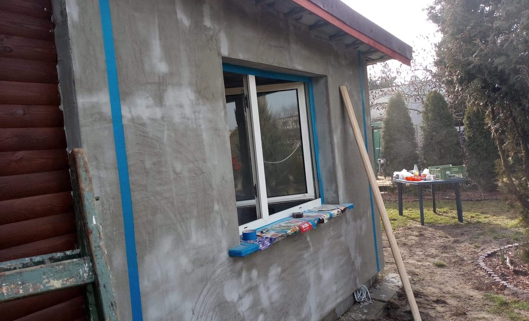 Ściana budynku w trakcie przygotowania do prac elewacyjnych: surowy tynk, zabezpieczone okno folią i taśmą malarską, widoczne ślady po wcześniejszych pracach, na parapecie leżą narzędzia...
