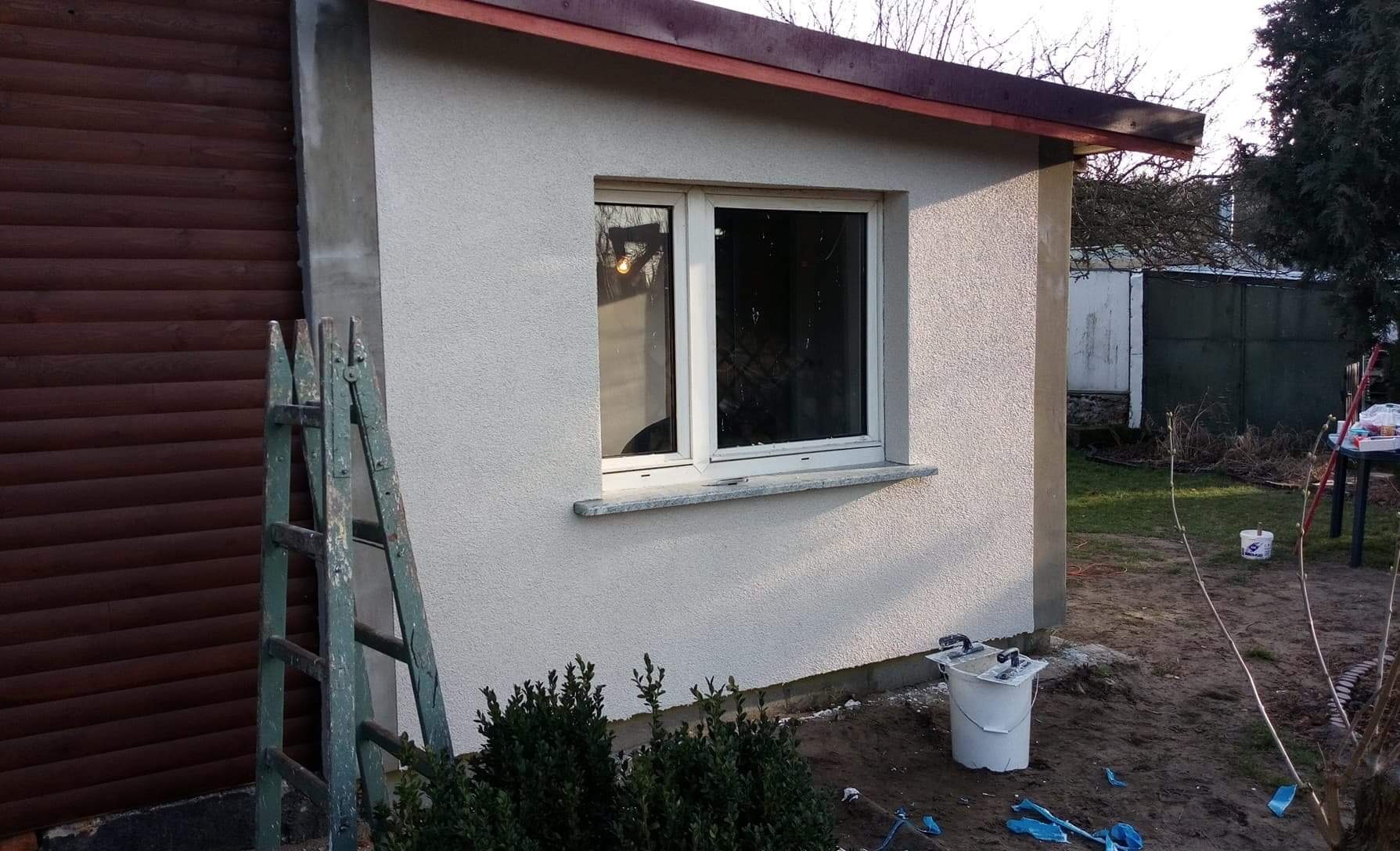 Ściana budynku po nałożeniu nowej, jasnoszarej struktury tynku, widoczne okno z białymi ramami i kamiennym parapetem, obok drewniana drabina oparta o ścianę i wiadro z resztkami materiałów budowlanych