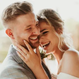 Uśmiechnięta para młoda w objęciach, panna młoda z dłonią na twarzy pana młodego, widoczny pierścionek zaręczynowy i butonierka.