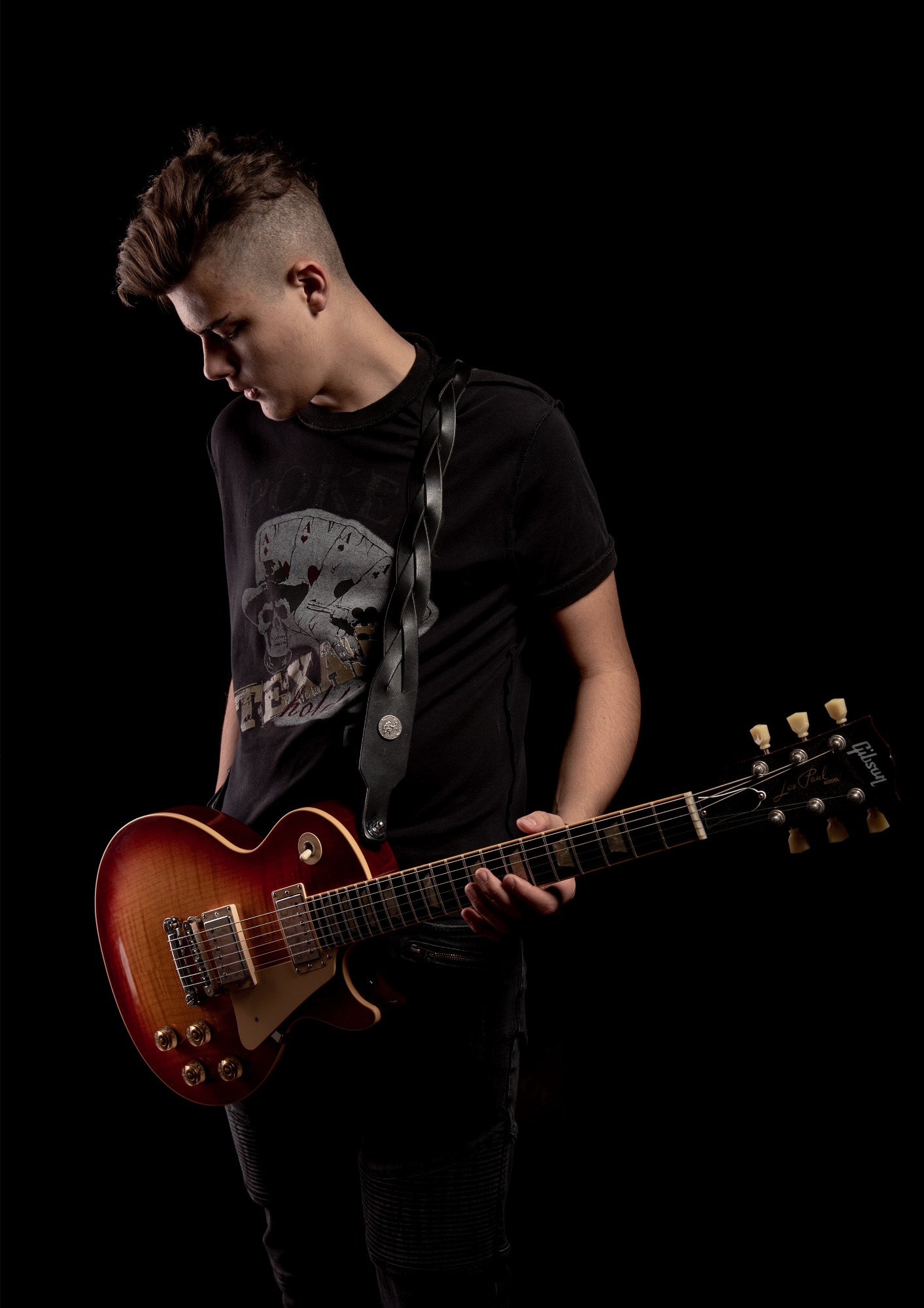 Młody mężczyzna z irokezem, w czarnym t-shircie z nadrukiem kart i czaszki, trzymający gitarę elektryczną Gibson Les Paul z brązowo-czerwonym wykończeniem, na czarnym tle.