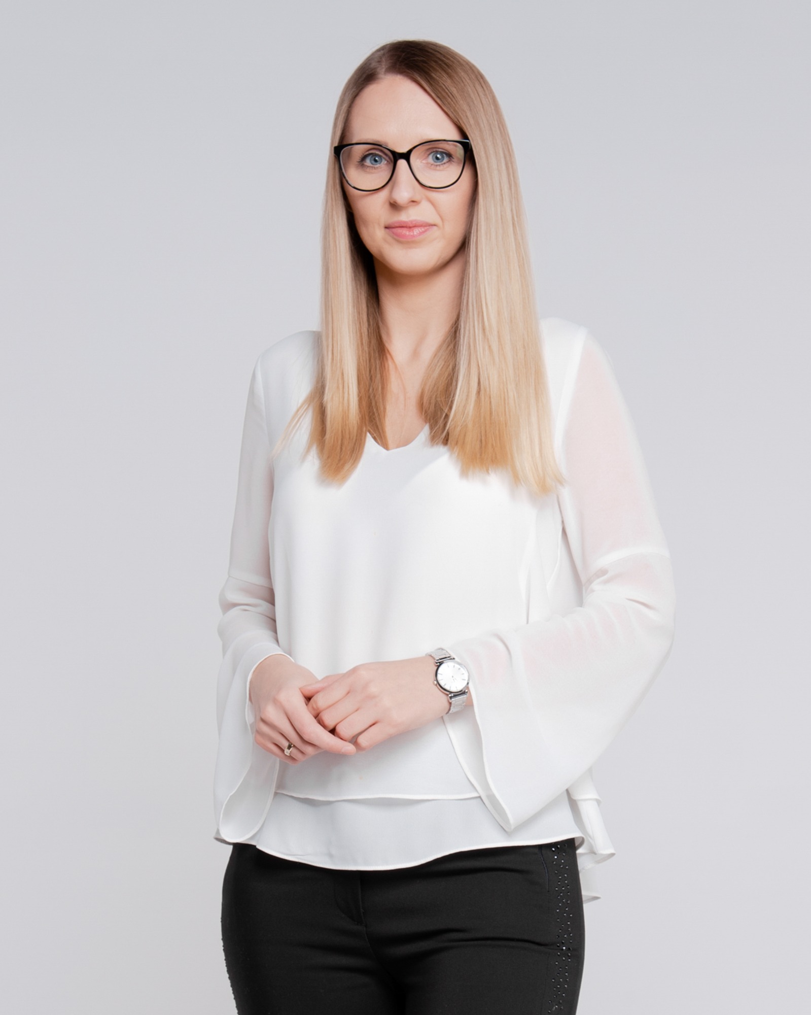 Portret kobiety w okularach, białej bluzce z szerokimi rękawami i czarnych spodniach, na jasnym tle, z delikatnym uśmiechem i splecionymi dłońmi.