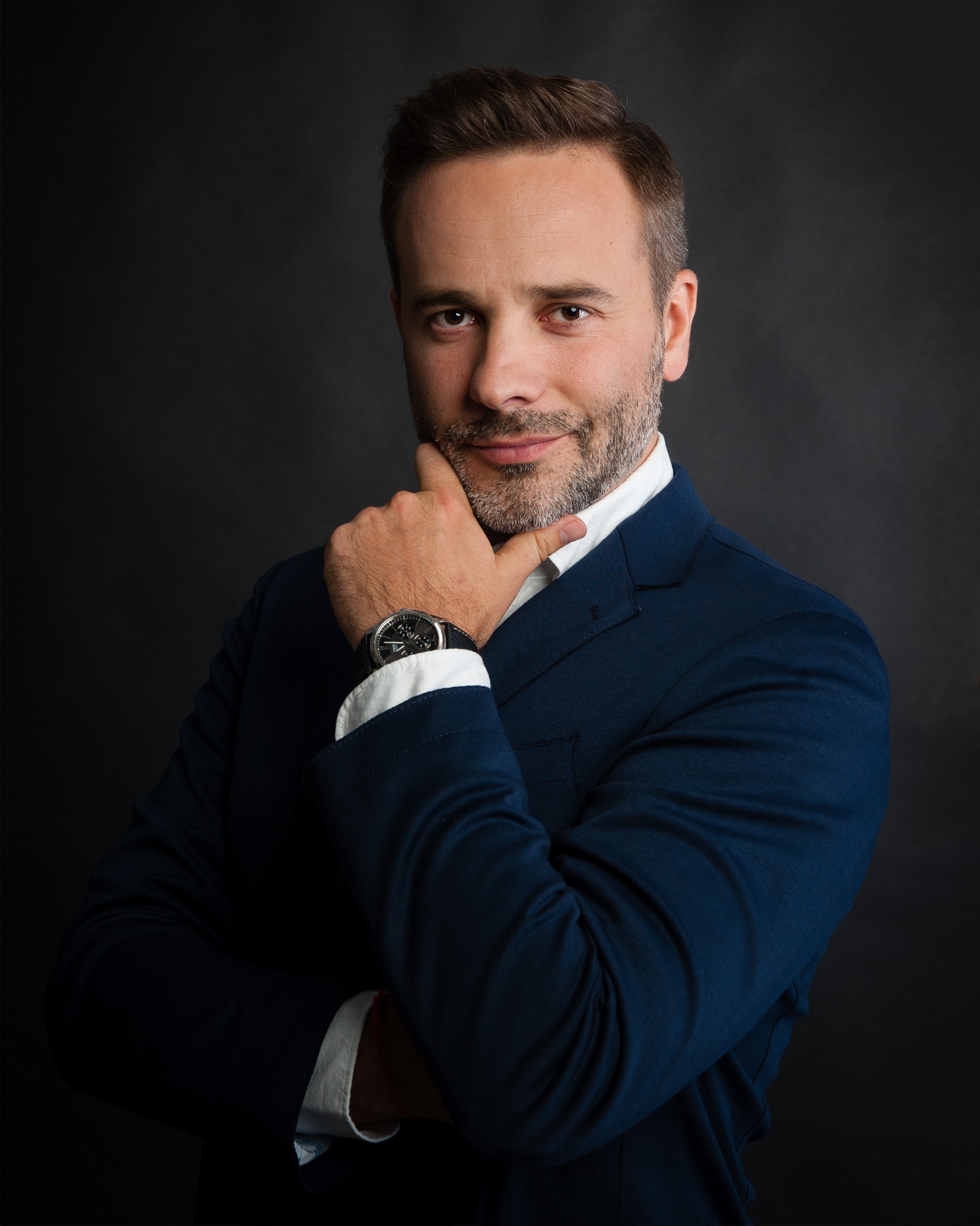 Portret mężczyzny w niebieskiej marynarce, z ręką pod brodą, prezentującego zegarek na tle ciemnego tła.