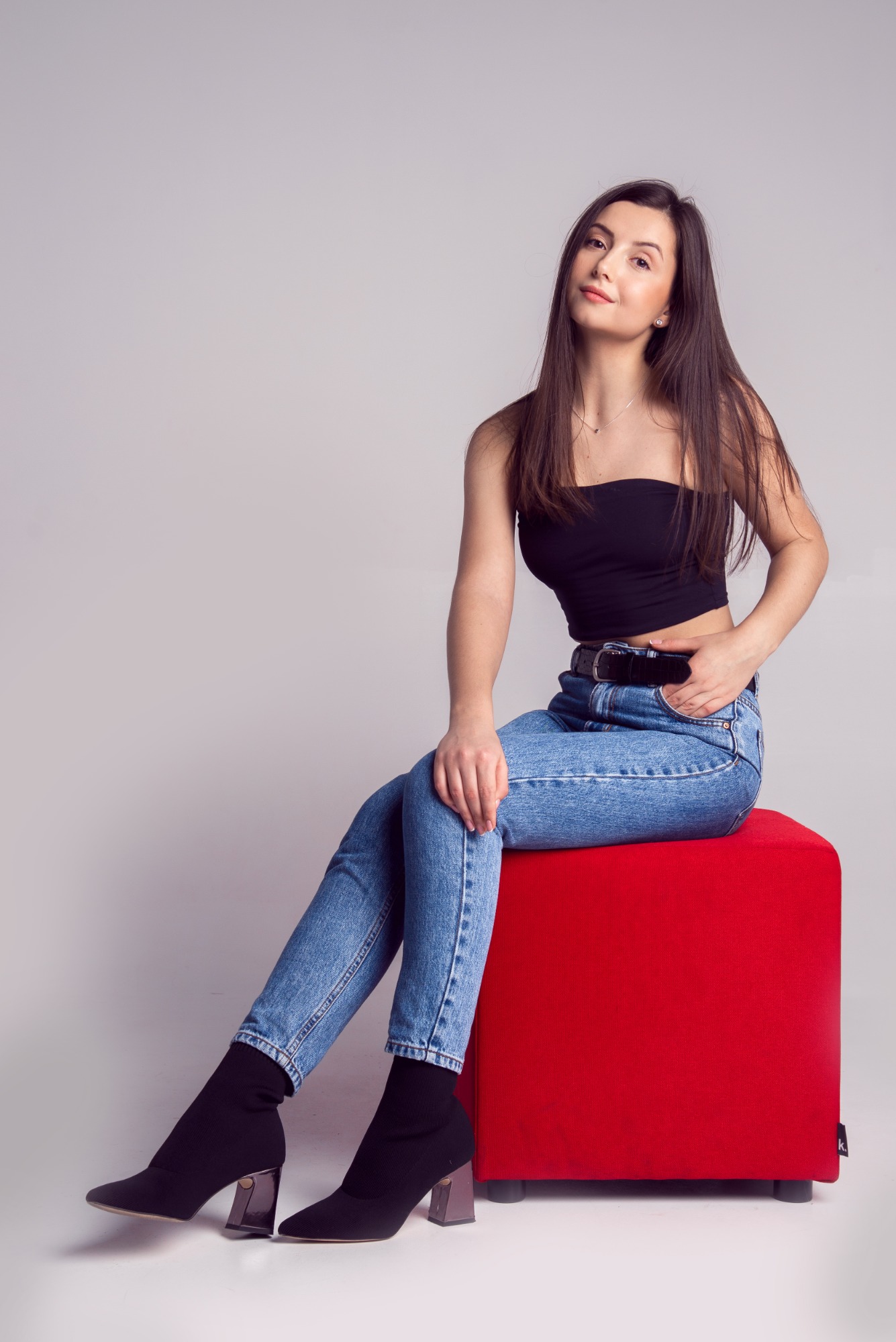 Portret kobiety siedzącej na czerwonym pufie, ubrana w dżinsy, czarny top i botki na obcasie, z ręką w kieszeni i delikatnym uśmiechem.